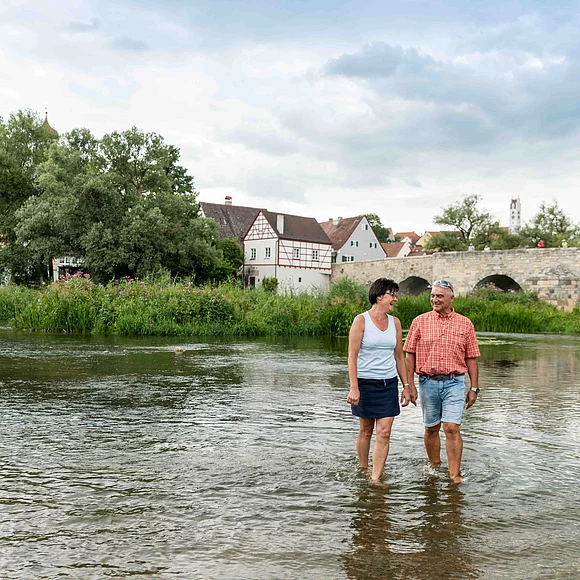 Ein Paar watet am Wörnitzstrand durchs Wasser. Im Hintergrund sieht man die steinerne Brücke und Häuser von Harburg.