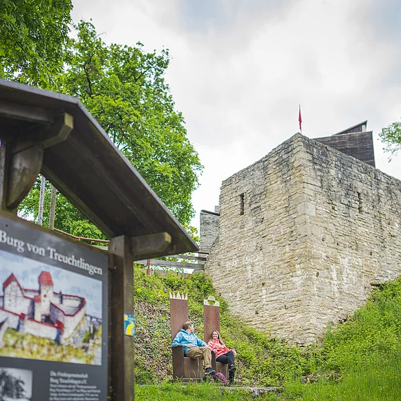 im Vordergrund groß die Schautafel zur Oberen Burg, ein junges Paar sitzt auf den Thronen mittig im Bild vor der Burgkulisse