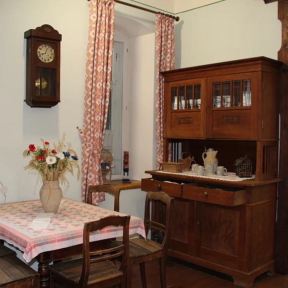 Eine Wohnstube mit großem Schrank, einem Tisch mit Stühlen und eine über dem Tisch an der Wand angebrachte hölzerne Uhr