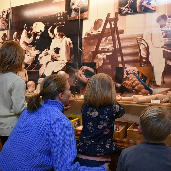 Eine Frau sieht sich mit drei Kindern eine Vitrine im Käthe-Kruse-Puppenmuseum an in der erklärt wird, wie die Puppen hergestellt werden.