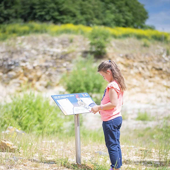 In einer flachen Grube aus Lockergestein mit vereinzelter Vegetation betrachtet ein Mädchen eine Informationstafel für Kinder.