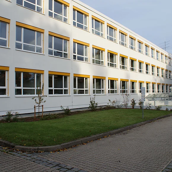 Ein langes dreistöckiges Gebäude hat eine weiße Fassade, große Fenster und gelbe Rolladenkästen.
