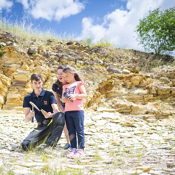 Vor einer Felswand aus plattigem Kalkgestein zeigt ein Mann mit Geologenhammer einer Frau und einem Mädchen ein Gesteinsbruchstück aus der Felswand. Der Boden ist ebenfalls mit Bruchstücken von Kalkgesteinsplatten bedeckt.