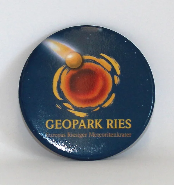 Ein runder Magnet mit Flaschenöffner auf der Rückseite zeigt das Logo des Geopark Ries auf dunkelblauem Hintergrund.