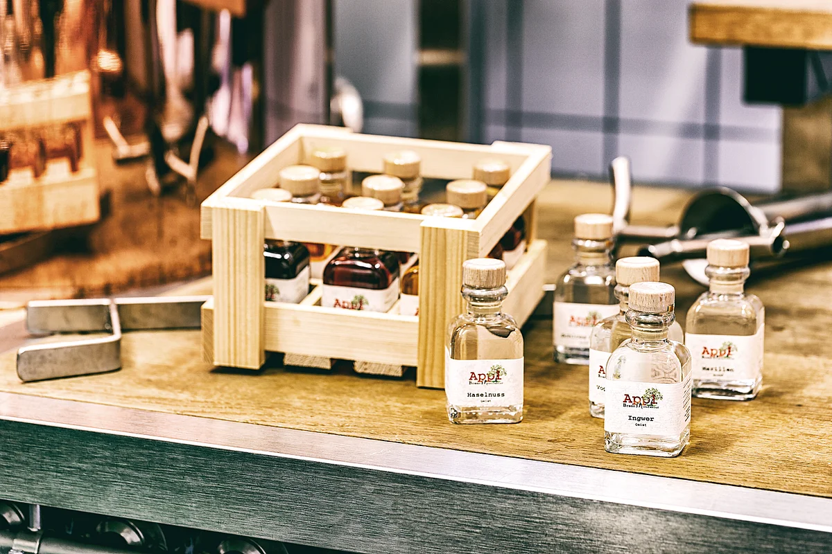 In einer kleinen Holzkiste und auf Tisch daneben stehen kleine Glasfalschen mit Holzkorken und der Aufschrift "Appl" auf dem Etikett.