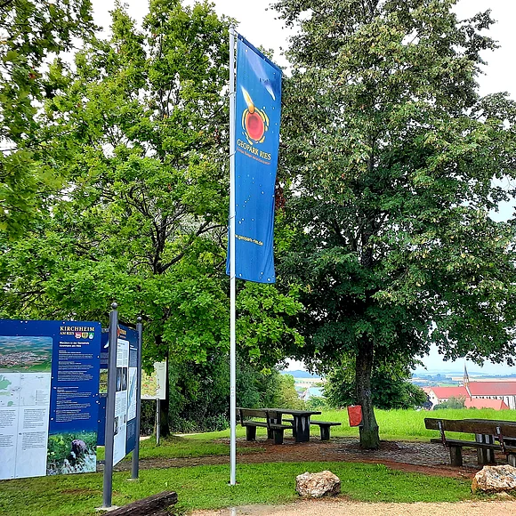 Unter hohen Bäumen stehen zwei Sitzbänke und daneben Informationstafeln und eine Fahne des Geopark Ries. Im Tal ist eine Kirche mit kleinem Turm zu sehen.
