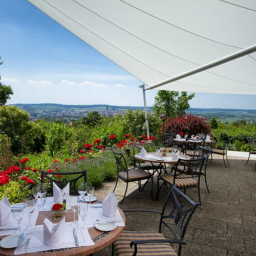 Auf einer von roten Rosen umgebenen Terrasse sind viele festlich gedeckte Tische. In der Senke im Hintergrund ist eine Stadt zu sehen.