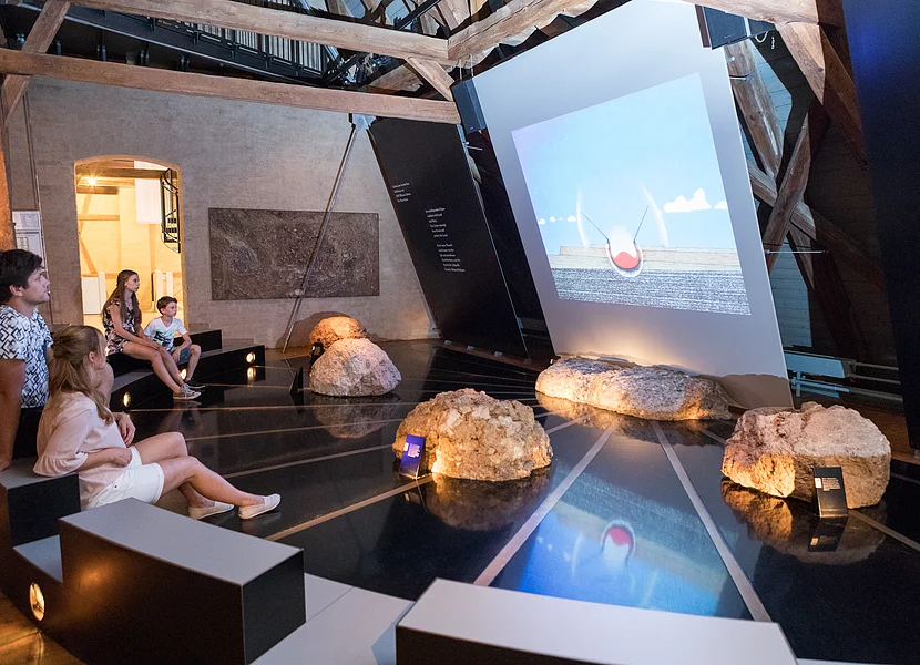 Eine Familie sitzt auf halbrund angeordeten Sitzbänken vor einer Leinwand, die eine Animation eines Meteoriteneinschlags zeigt. Vor der Leinwand liegen einige Felsblöcke.