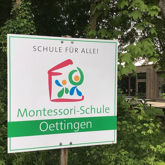 Das Bild zeigt ein Schild mit dem Text "Schule für Alle - Montessori-Schule Oettingen"