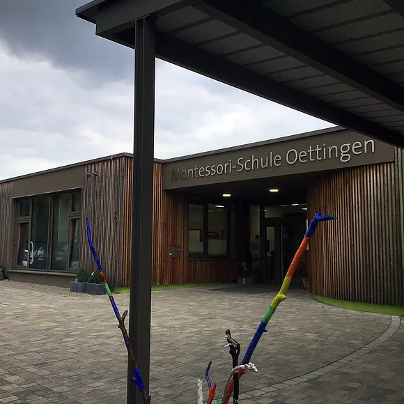 Seitlicher Blick auf den Eingang der Montessori-Schule Oettingen. Im Vordergrund ein bunt bemalter Ast.