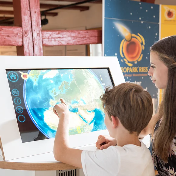 Zwei Kinder stehen vor einem Monitor mit einem Satellitenbild der Erde. Im Hintergrund sind Informationstafeln mit dem Logo des Geopark Ries zu sehen.