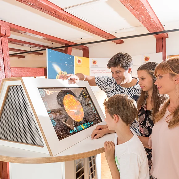 Eine Familie steht vor einem Monitor, der einen Planeten zeigt. Durch den Raum und an der Decke verlaufen rote Holzbalken.