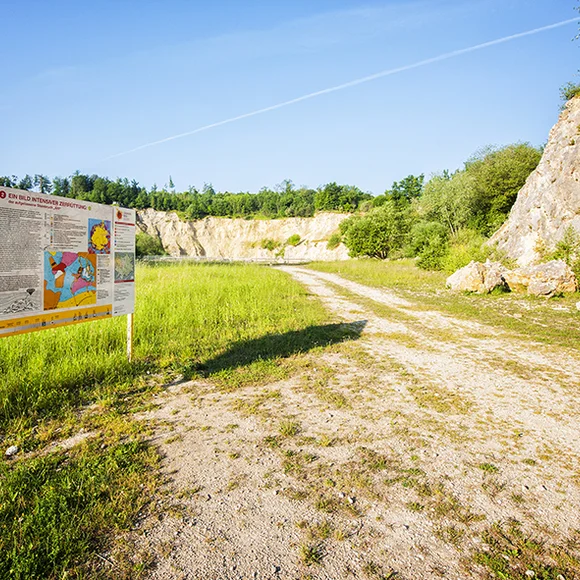 Neben einem Schotterweg, der in einen ehemaligen Steinbruch führt, steht eine große Informationstafel.