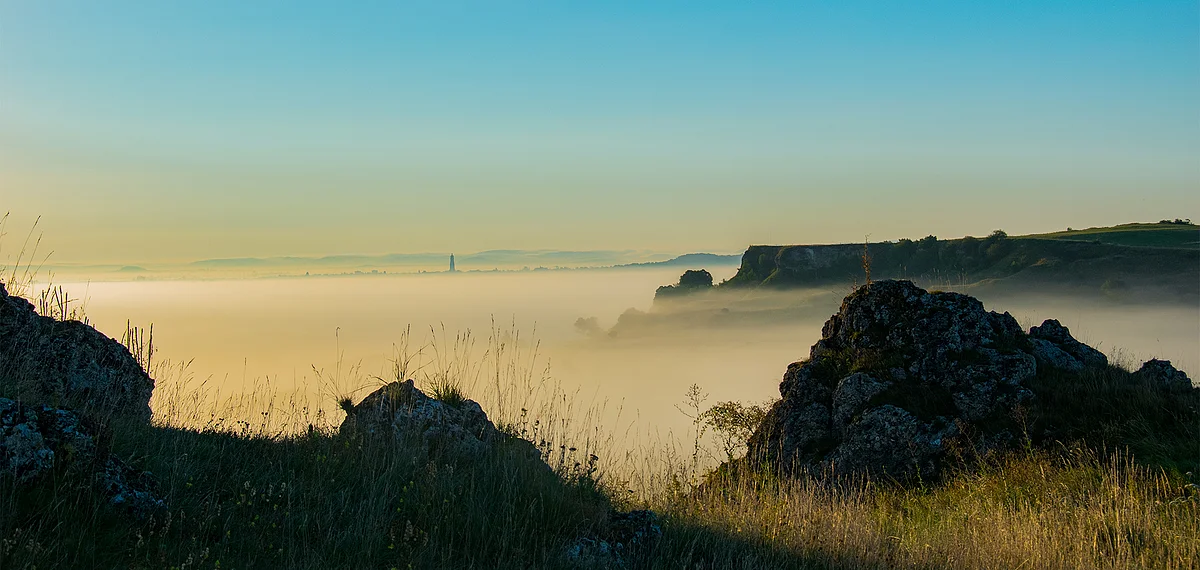 Aus einer dichten Nebeldecke ragt ein plateauförmiger Hügel und ein Kirchturm. Im Vordergrund sind schemenhaft einzelne Felsblöcke zu sehen.