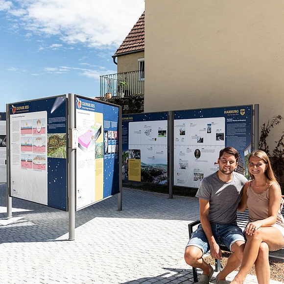 Ein Mann und eine Frau sitzen auf einer Bank bei mehreren Informationstafeln über den Geopark Ries.