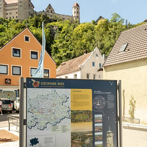 Unterhalb einer Burg steht zwischen Häusern eine Informationstafel über den Geopark Ries.