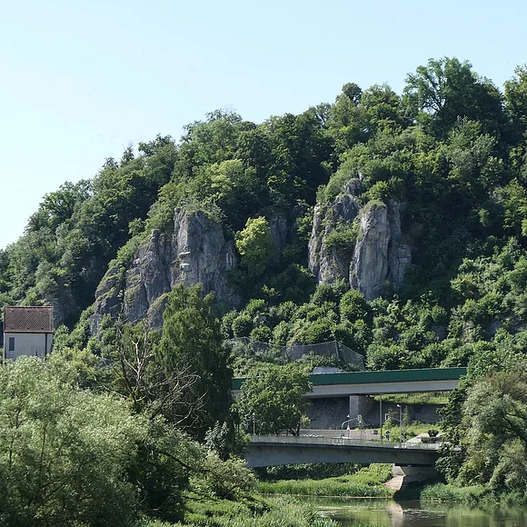 An einem mit Bäumen bewachsenen Hang befinden sich zwei rundliche Felsformationen. Im Vordergrund sind zwei Brücken und ein Fluss zu sehen.