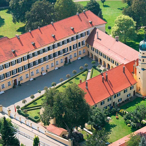 Die Luftaufnahme zeigt mehrere aneinander angrenzende Gebäude mit gelber Fassade und grünen Fensterläden. Die angrenzenden Grünanlagen wirken gepflegt.