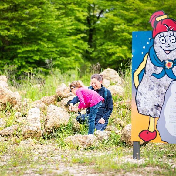 Neben einer Informationstafel mit der Figur Suevitchen klopfen ein Mädchen und eine Frau Steine.