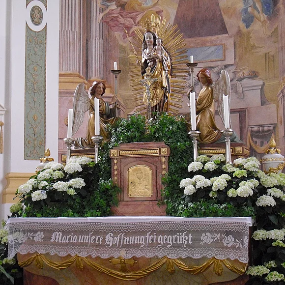 Der mit weißen Blumen geschmückte Marienaltar. In der Mitte die Marienstatue mit Kind. Zu ihren Füßen knien beidseitig Engel mit Kerzen.