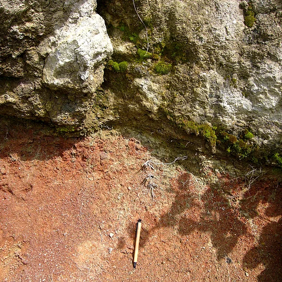 Unterhalb eines Felsblocks befindet sich sehr feines, rötliches Lockermaterial. Zum Größenvergleich liegt dort ein Stift.