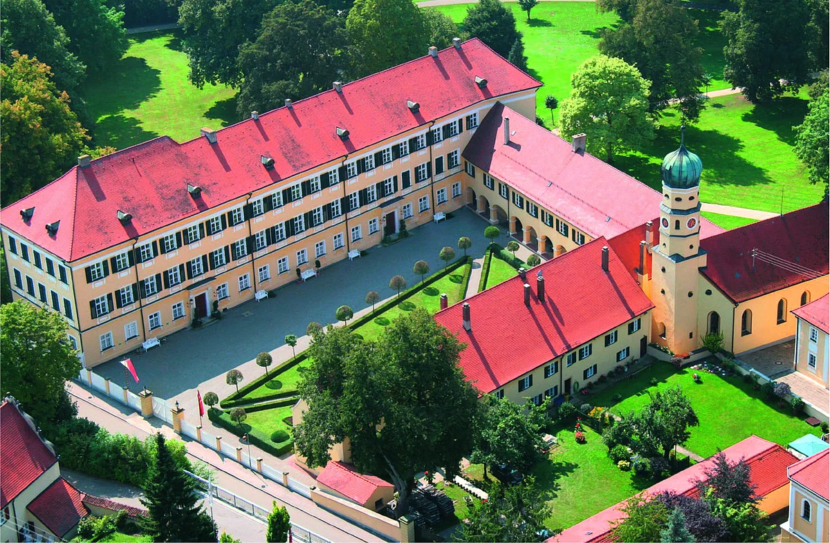 Die Luftaufnahme zeigt mehrere aneinander angrenzende Gebäude mit gelber Fassade und grünen Fensterläden. Die angrenzenden Grünanlagen wirken gepflegt.