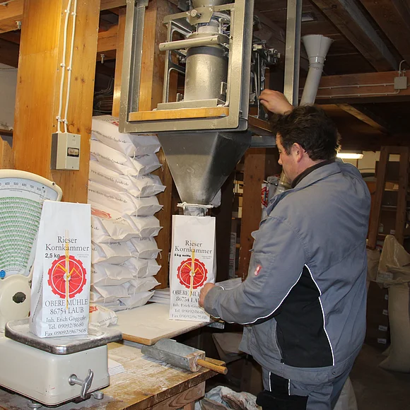 Ein Mann befüllt eine Mehltüte mit der Aufschrift "Rieser Kornkammer". Auf der Waage daneben steht eine bereits befüllte Tüte.