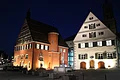 Neben einem beleuchteten Haus mit roter Fassade steht ein weiteres Haus mit weißer Fassade. Der Himmel ist dunkel.