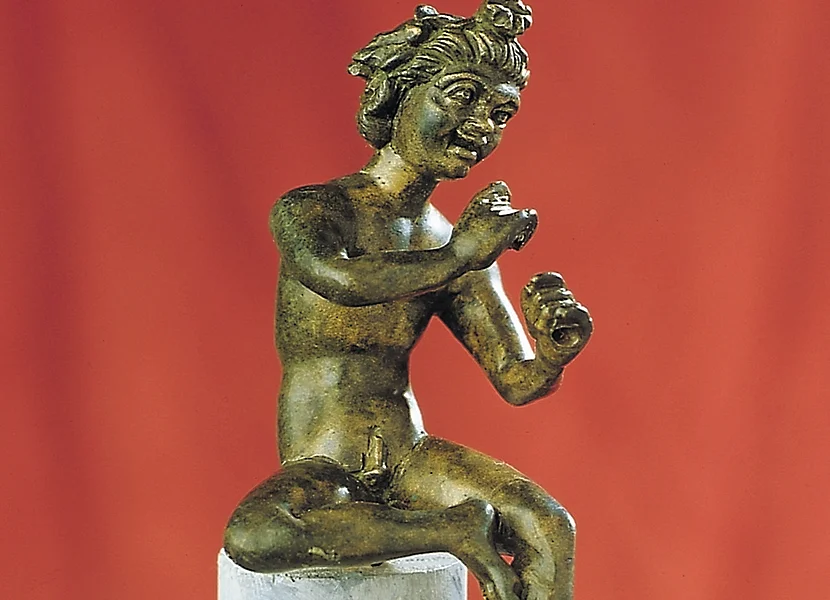 Auf einem kleinen Stab sitzt eine Bronzefigur, die die Hände wie zum Flötenspiel hält.