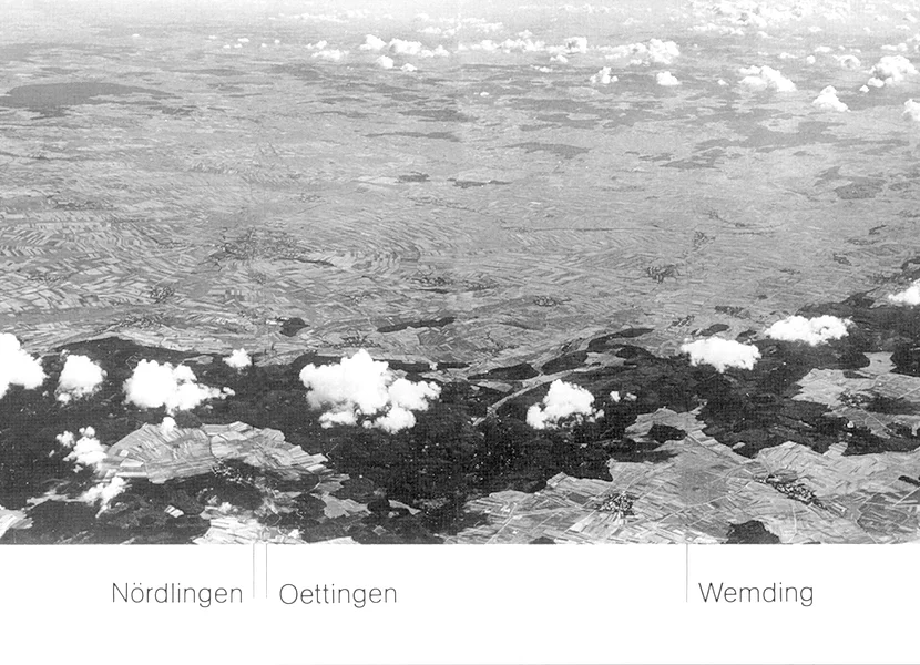 Die schwarz-weiße Luftaufnahme zeigt den Rieskrater mit dem bewaldeten Kraterrand und Wolken darüber. Linien zeigen die Lage der Städte Nördlingen, Oettingen und Wemding.