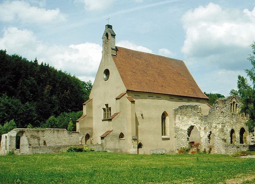 An eine kleine braune Kirche schließen sich teils verfallene Mauern mit Rundbögen an.