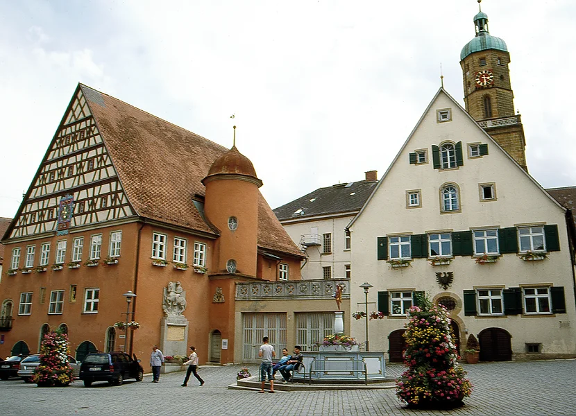 An einem gepflasterten Platz befinden sich ein rotes Gebäude mit Fachwerk am Giebel und ein cremefarbenes Gebäude mit Fensterläden, hinter dem sich ein Kirchturm erhebt.