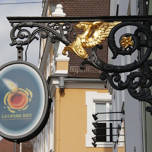 Ein gusseisernes verschnörkeltes Aushängeschild hat in der Mitte eine goldene Tierfigur mit Flügeln. In einem Rahmen ist das Logo des Geopark Ries abgebildet.