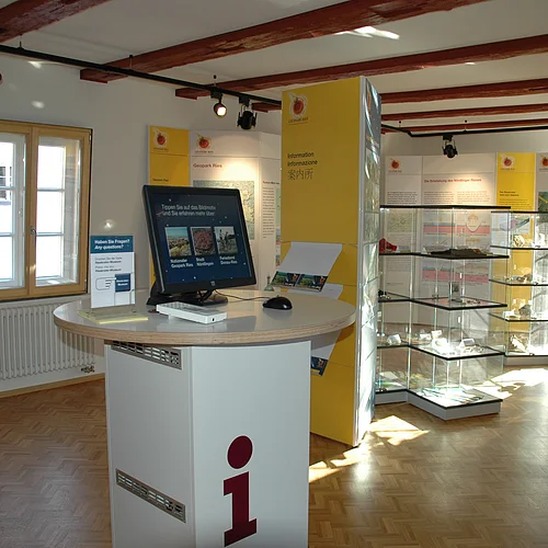 In einem Ausstellungsraum stehen mehrere Vitrinen und große Informationstafeln. Im Vordergrund steht auf einer Theke ein Computermonitor.