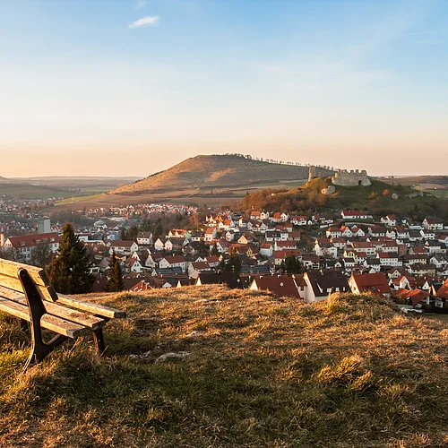 Im Vordergrund steht eine von der Abendsonne beschienene Holzbank auf einem Hügel. In einiger Entfernung erhebt sich die Ruine Flochberg und der Ipf, im Tal liegt die Stadt Bopfingen.