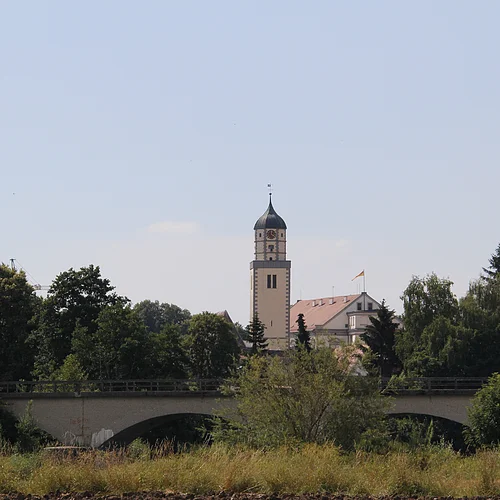 Hinter Bäumen erhebt sich ein cremefarbener Kirchturm. Im Vordergrund ist eine Brücke mit Bögen zu sehen.