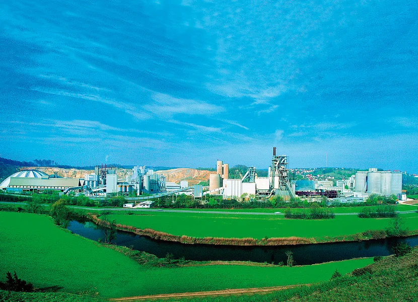 In einer Ebene steht die große Produktionsanlage eines Zementwerks mit vielen Silos. Im Vordergrund schlängelt sich zwischen zwei Wiesen ein Fluss.