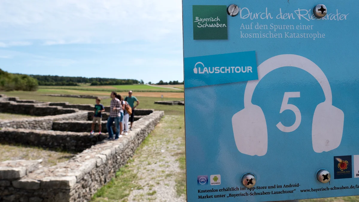 Ein kleines Schild weist auf die Lauschtour 5 "Durch den Rieskrater" hin. Im Hintergrund sind Personen zu sehen, die auf den Mauerfundamenten eines Gebäudes laufen.