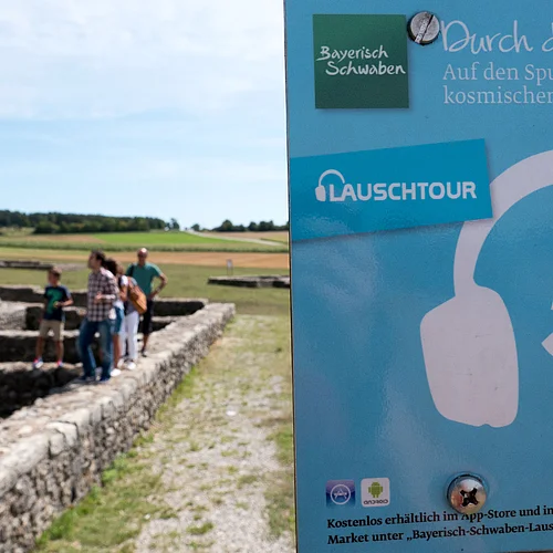 Ein kleines Schild weist auf die Lauschtour 5 "Durch den Rieskrater" hin. Im Hintergrund sind Personen zu sehen, die auf den Mauerfundamenten eines Gebäudes laufen.