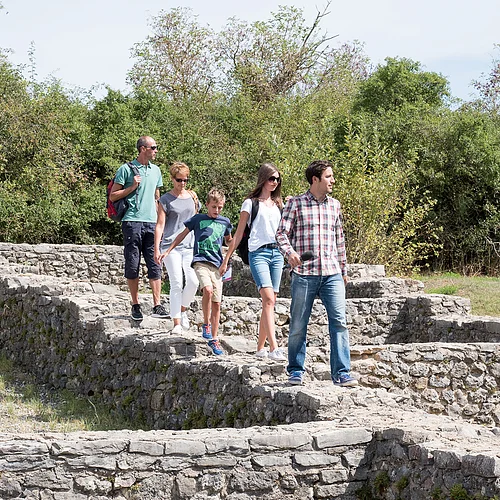 Fünf Personen laufen über die gemauerten Fundamente eines ehemaligen Gebäudes. Im Hintergrund ist eine Buschreihe zu sehen.