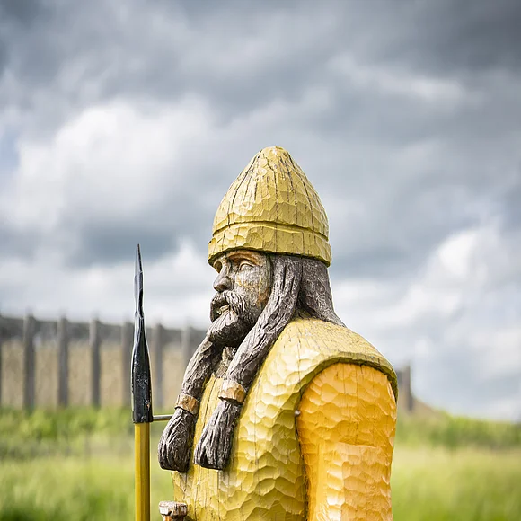 Auf einer Wiese steht die aus Holz geschnitzte und bemalte Figur eines bewaffneten keltischen Mannes. Im Hintergrund ist ein rekonstruiertes Bauwerk zu sehen.