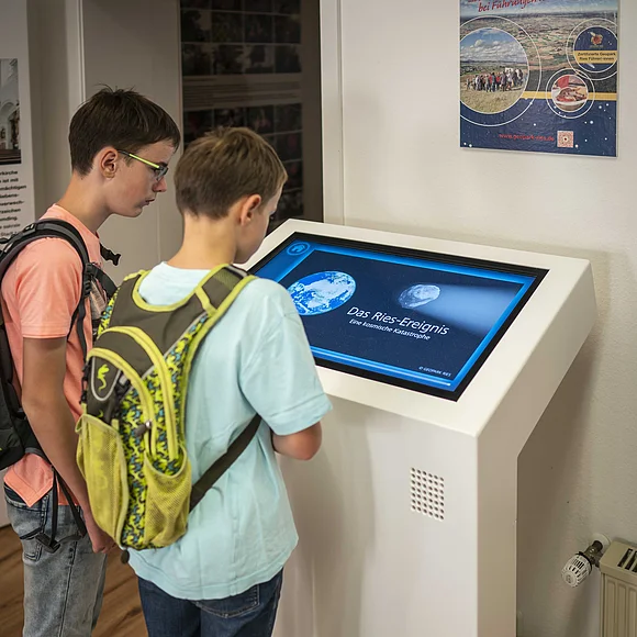 Zwei Jungen, die vor einem Bildschirm in der Geopark Ries Infostelle Wemding stehen, der eine Animation des Ries-Ereignisses zeigt.