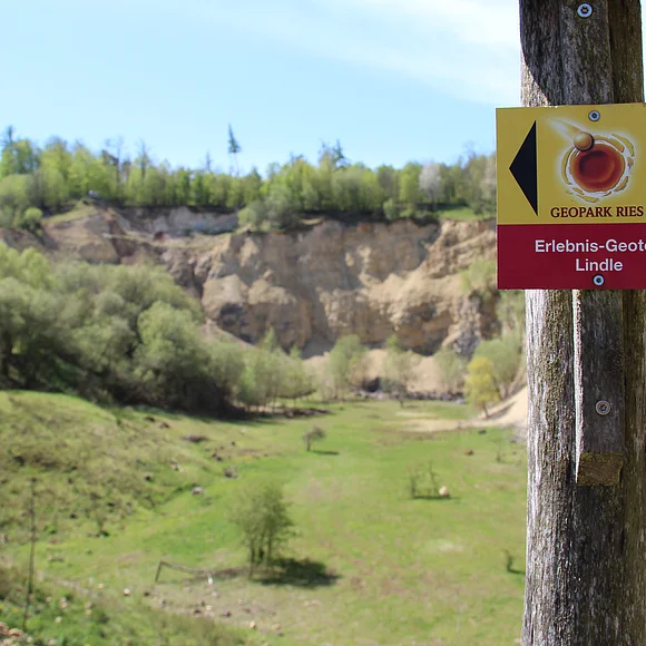 An einem Pfosten ist ein Wegweiserschild des Geopark Ries Erlebnis-Geotop Lindle angebracht. Im Hintergrund ist ein ehemaliger Steinbruch mit vielen Büschen und Bäumen zu sehen.