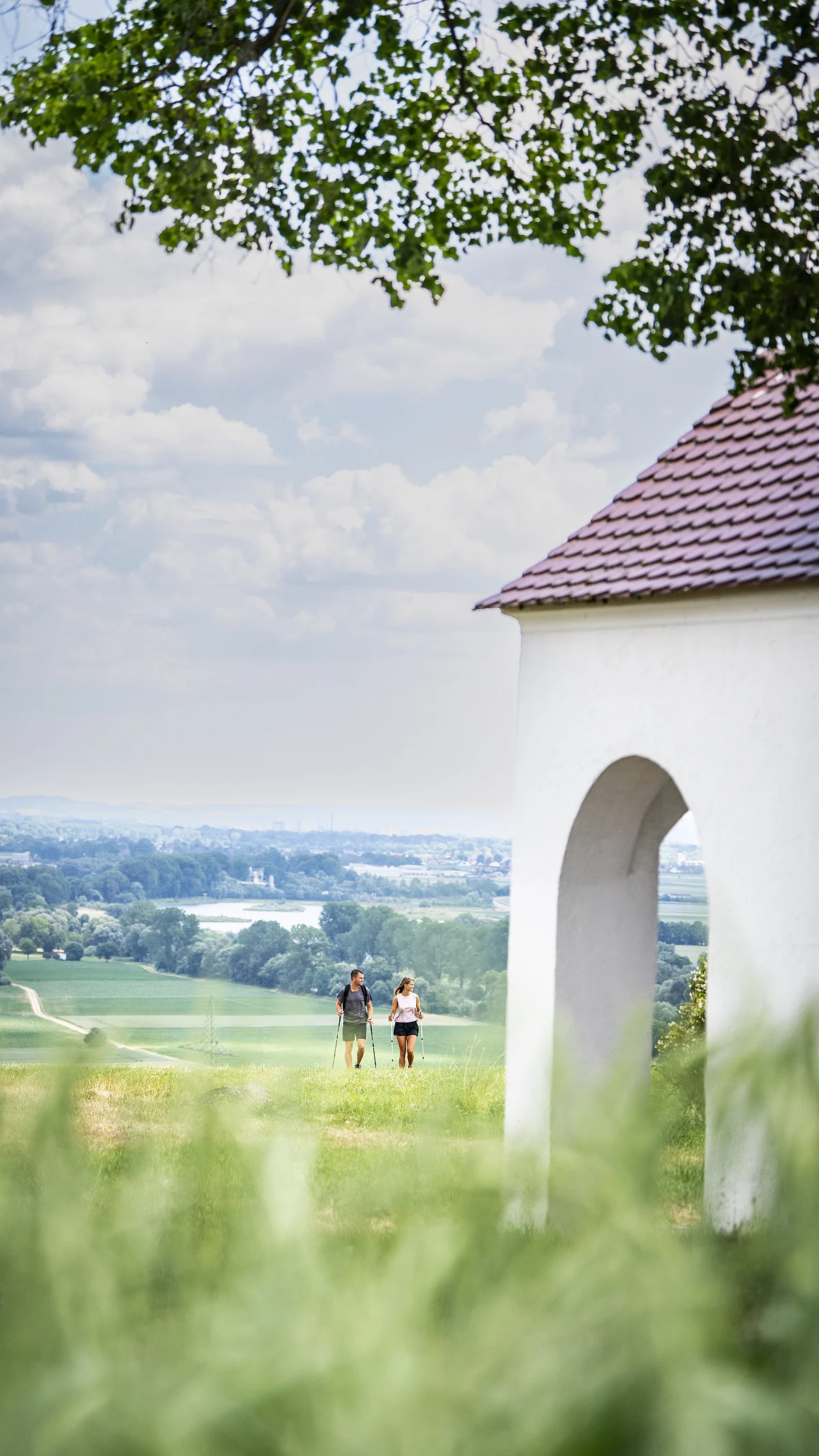 Zwei Personen wandern auf einem grünen Hügel, im Hintergrund eine weite Landschaft.