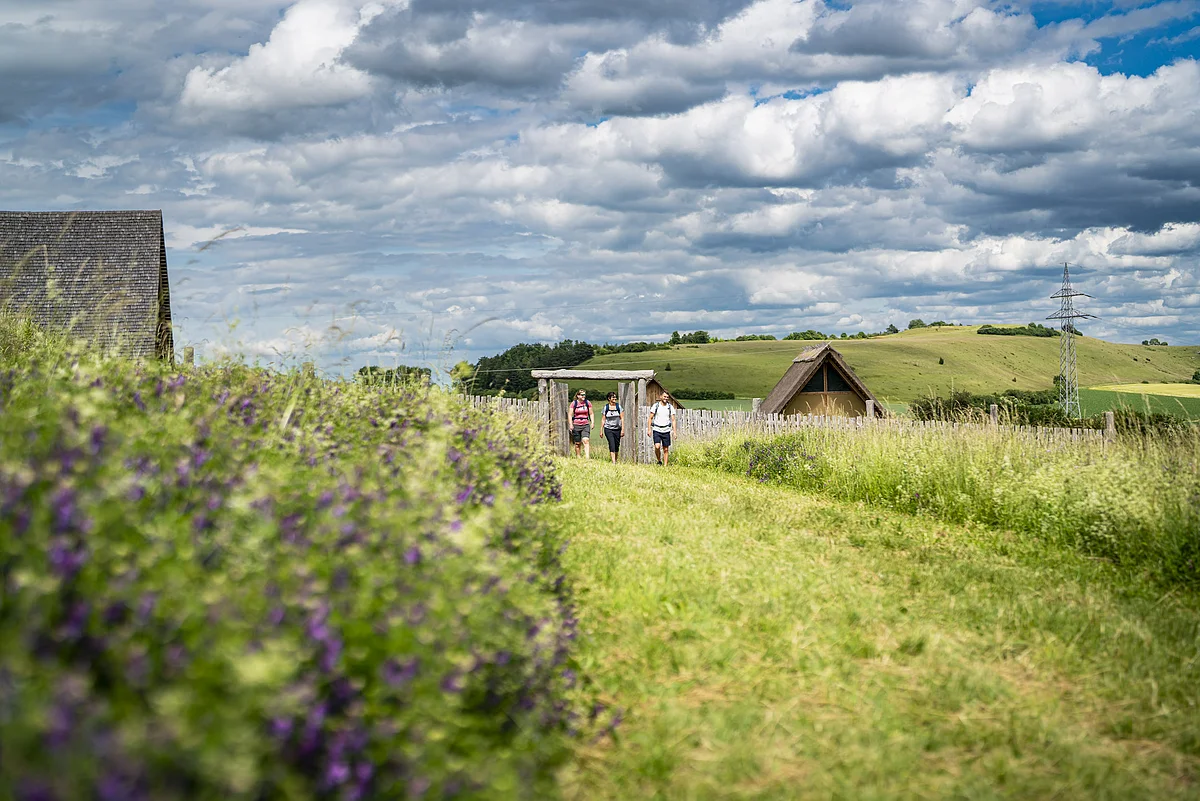 Drei Personen wandern auf einem Pfad durch eine ländliche Landschaft mit Hütten und blühenden Wiesen.