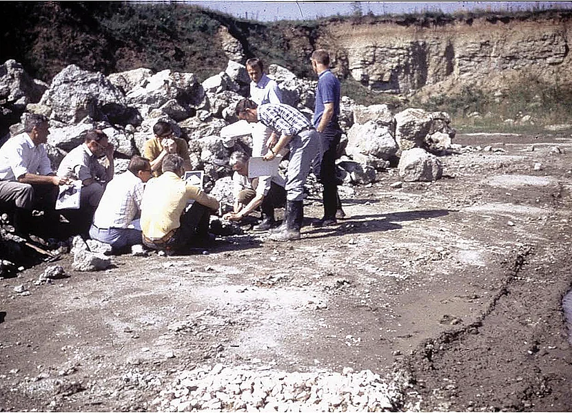 Mehrere Personen sitzen oder stehen neben einem Haufen Gesteinsbruchstücke. Im Hintergrund ist die Felswand eines ehemaligen Steinbruchs zu sehen.