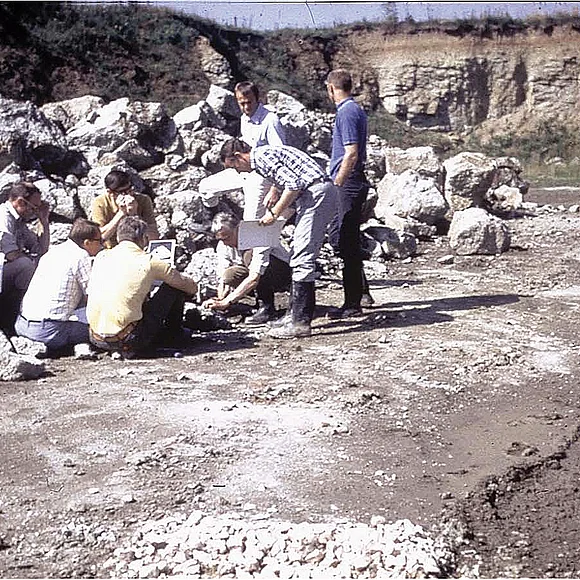 Mehrere Personen sitzen oder stehen neben einem Haufen Gesteinsbruchstücke. Im Hintergrund ist die Felswand eines ehemaligen Steinbruchs zu sehen.