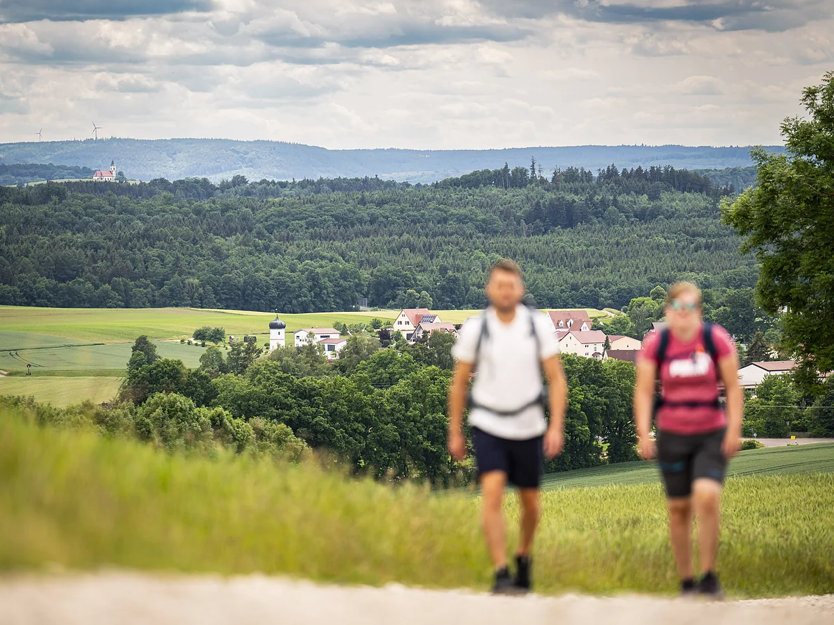 Zwei unscharfe Personen wandern auf einem Weg, im Hintergrund eine Landschaft mit Feldern, Wald und Gebäuden.