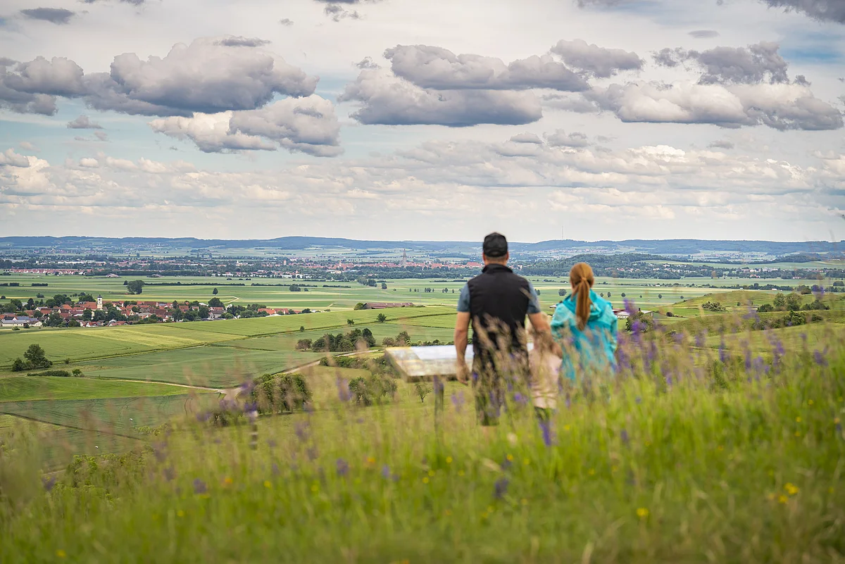 Zwei Personen stehen auf einem Hügel und blicken auf eine weite Landschaft mit Feldern und einem Dorf.