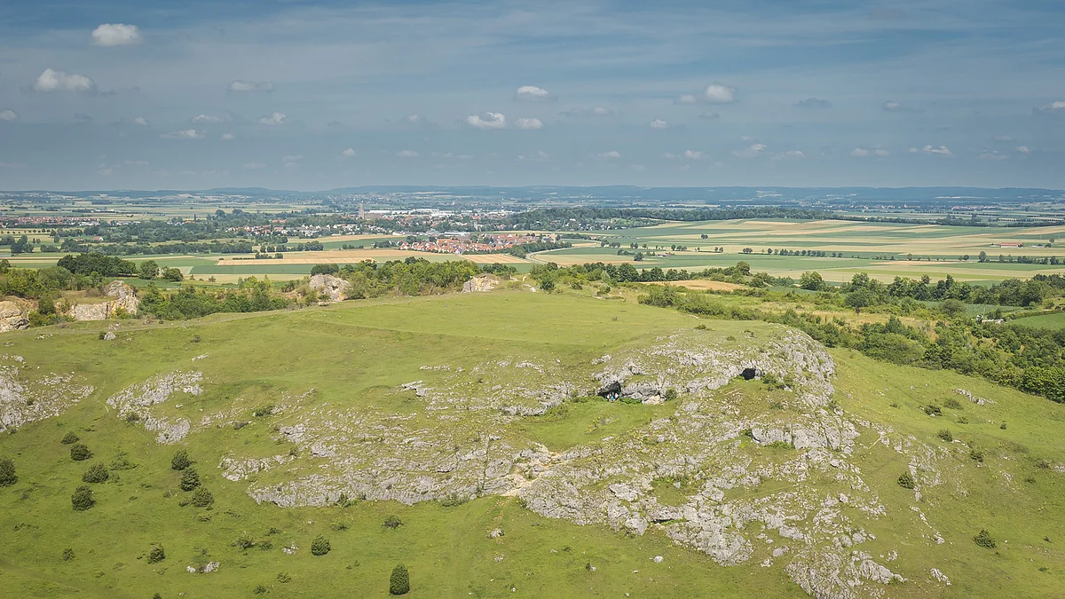 Hügelige Landschaft mit grünen Wiesen und Felsen im Vordergrund, Felder und ein Dorf im Hintergrund.